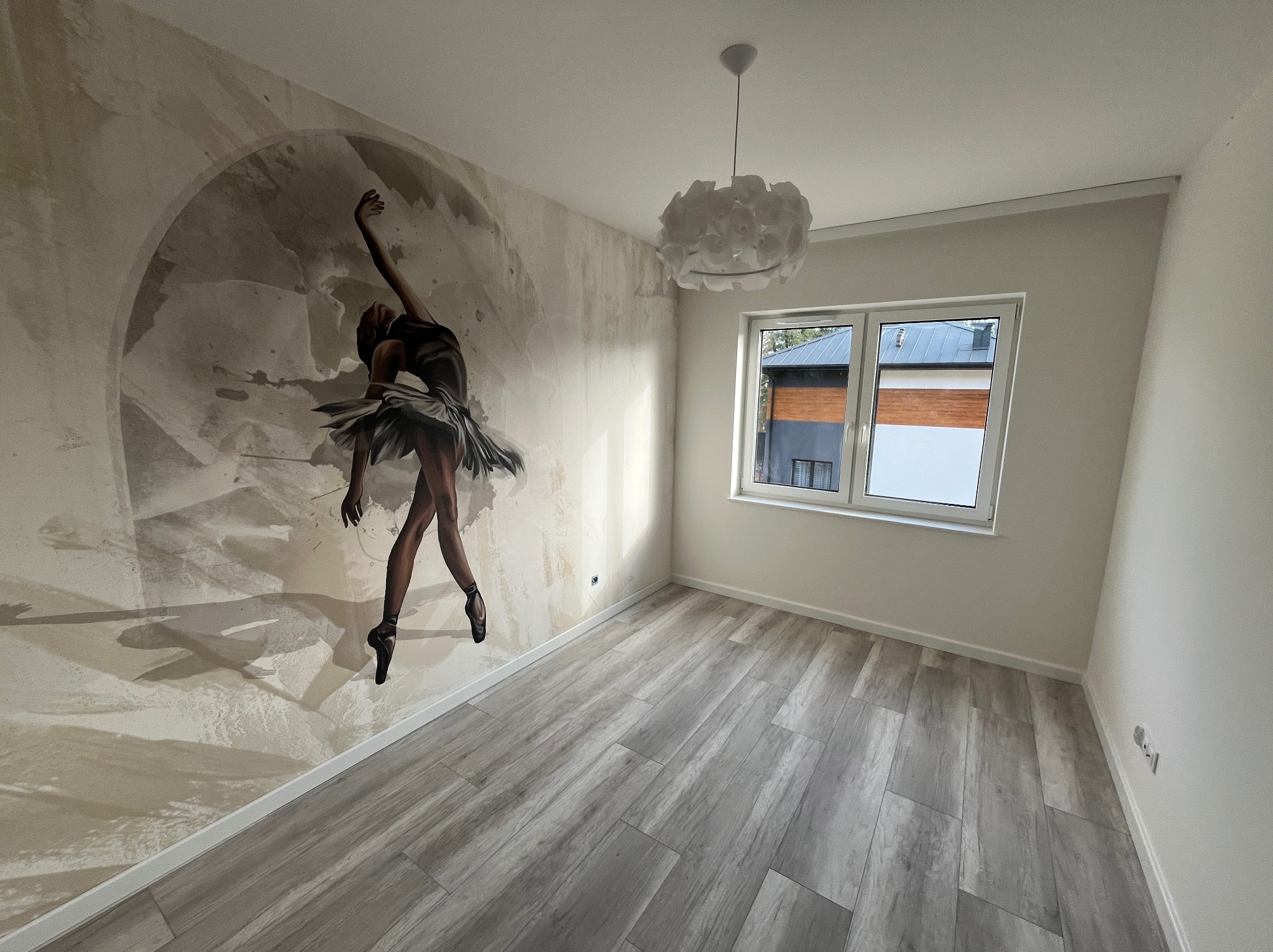 Pusty pokój po remoncie z szarymi panelami podłogowymi imitującymi drewno i fototapetą z motywem baletnicy na jednej ze ścian. Na suficie biała, nowoczesna lampa. Widoczne okno z widokiem na zewnątrz.