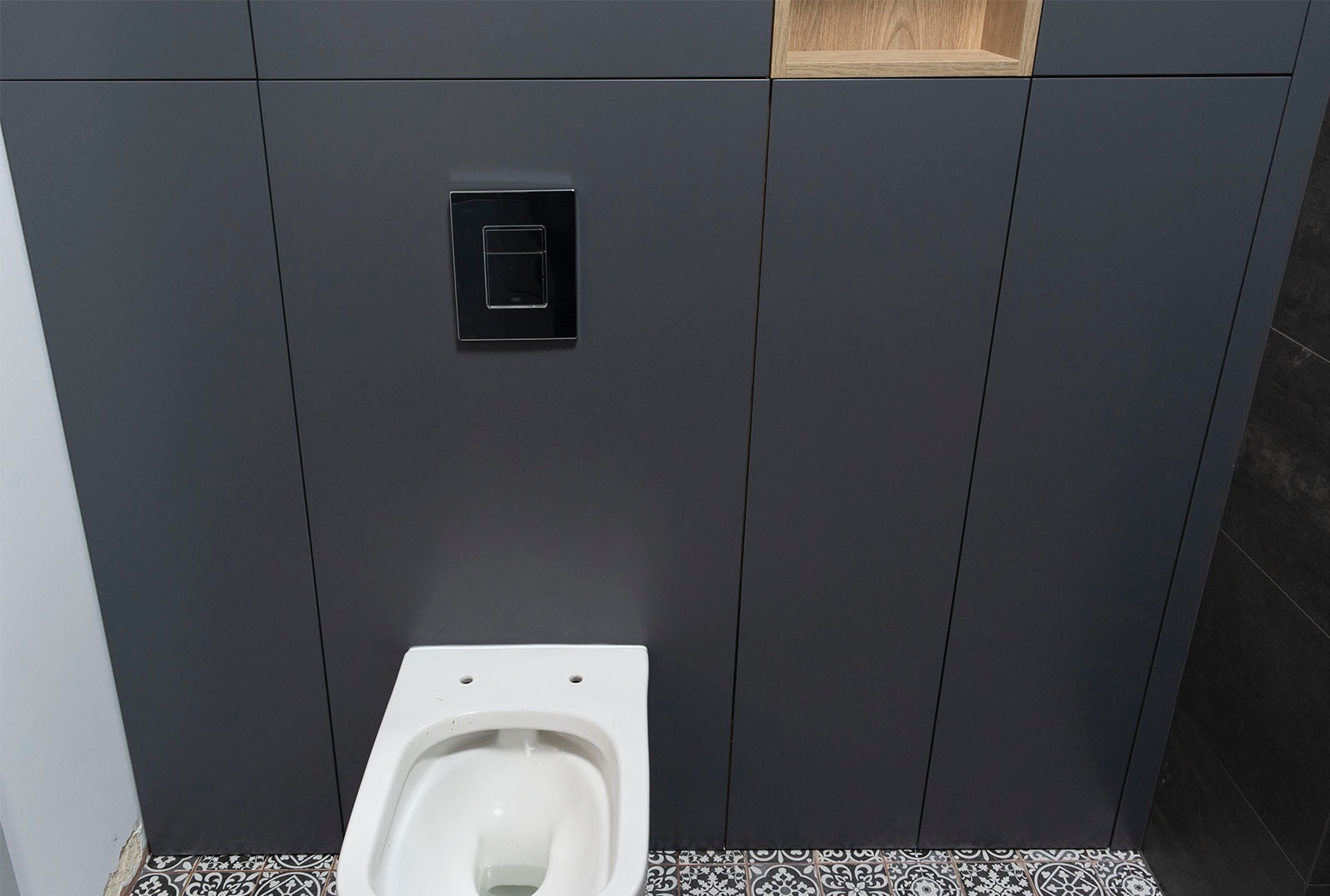 Wisząca toaleta z czarnym panelem spłukującym na tle grafitowych paneli ściennych z drewnianą wnęką, wzorzysta podłoga.