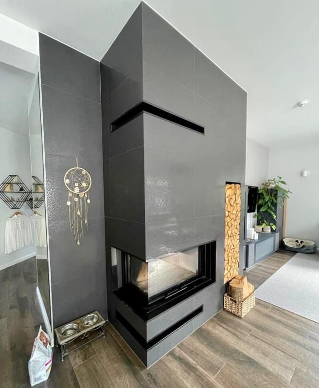 Nowoczesny kominek narożny obłożony szarą płytką ceramiczną z poziomymi wnękami, drewno ułożone pionowo obok, widok na wnętrze z dekoracjami i drewnianą podłogą.