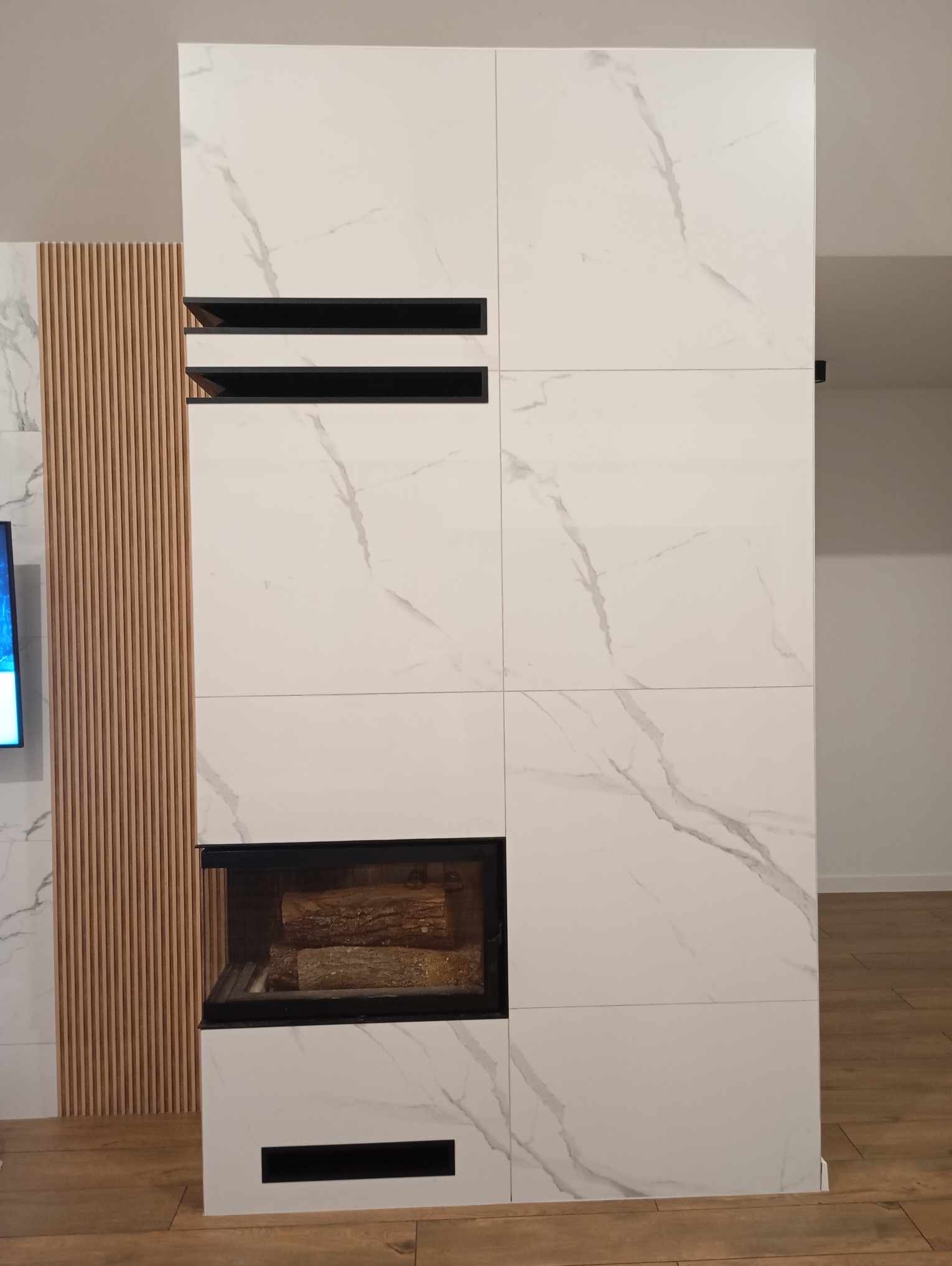 Nowoczesny kominek obudowany białymi płytami imitującymi marmur, z czarnymi wstawkami i drewnem w palenisku, sąsiadujący z dekoracyjną ścianką z drewnianych listewek.