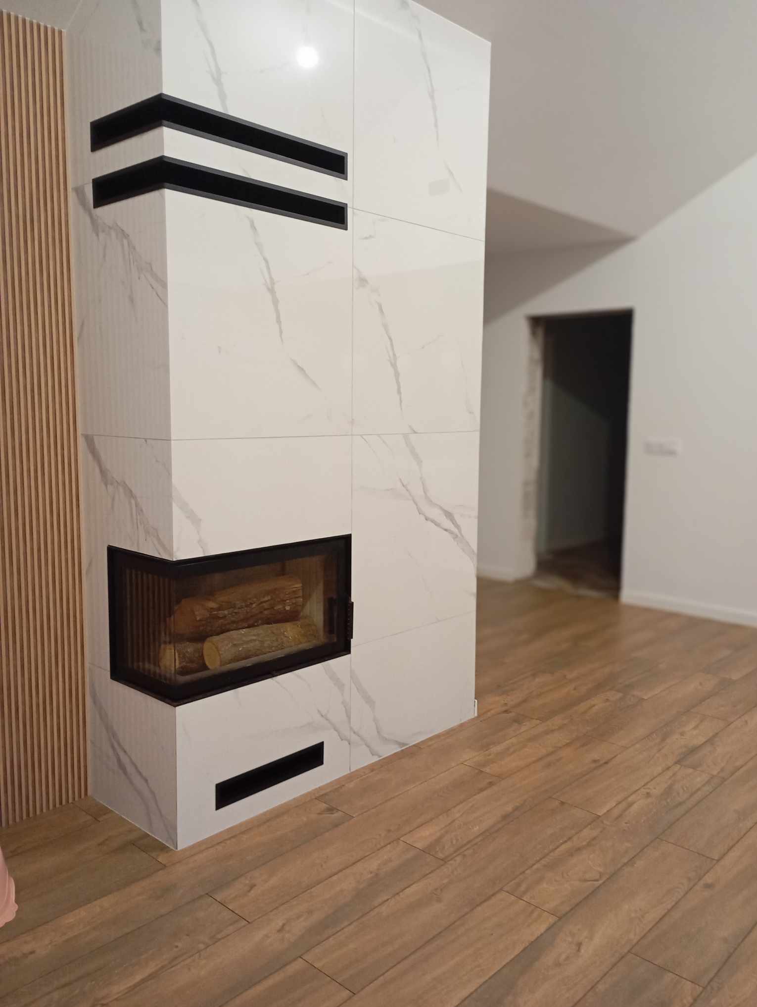 Obudowa kominka z białych płytek imitujących marmur, z czarnymi wstawkami i paleniskiem wypełnionym drewnem, na tle drewnianej podłogi i częściowo wykończonego wnętrza.