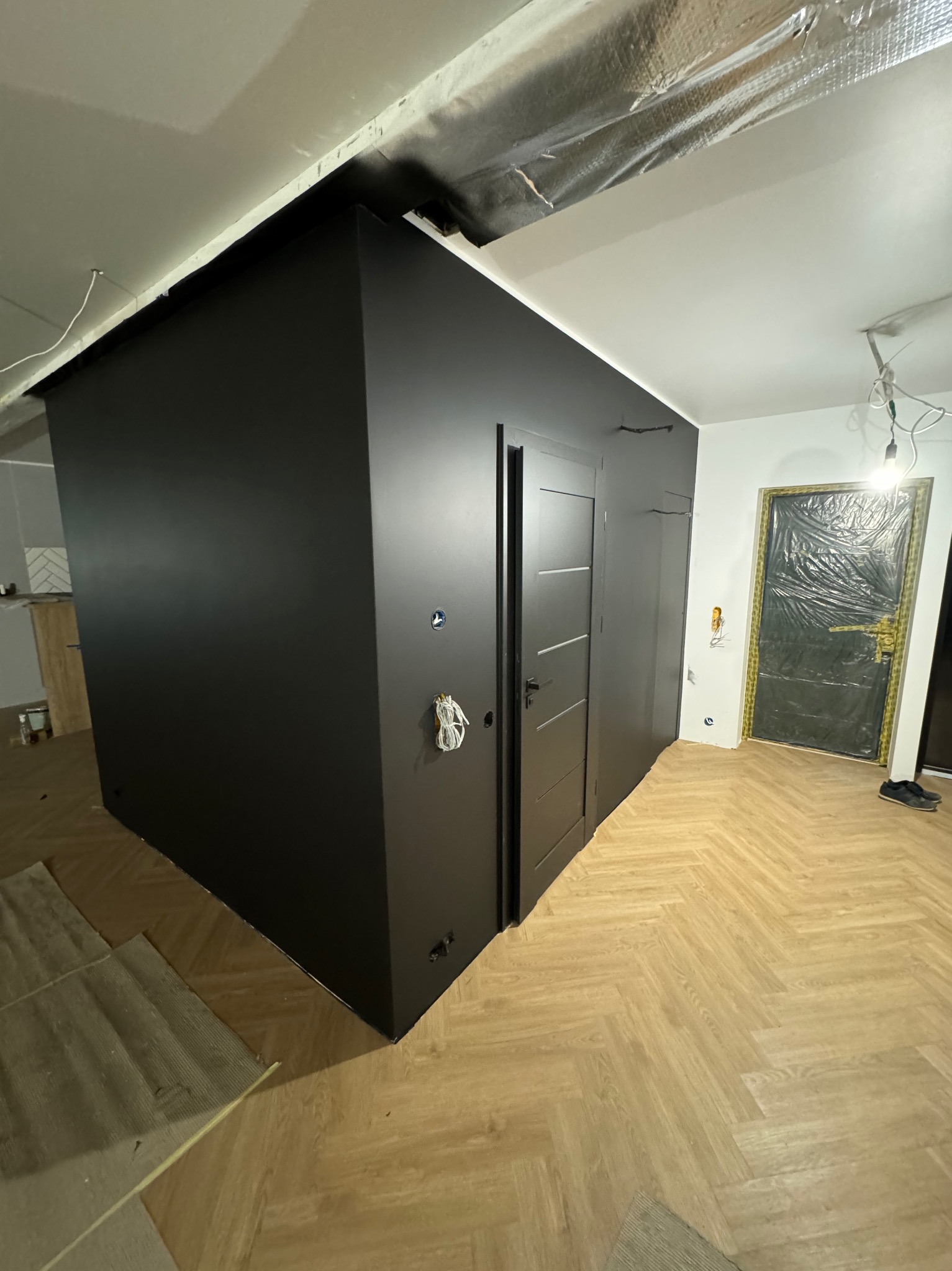 Ciemna zabudowa w trakcie wykańczania wnętrza: widoczne zabezpieczone drzwi folią, fragment instalacji elektrycznej i drewniana podłoga ułożona w jodełkę.