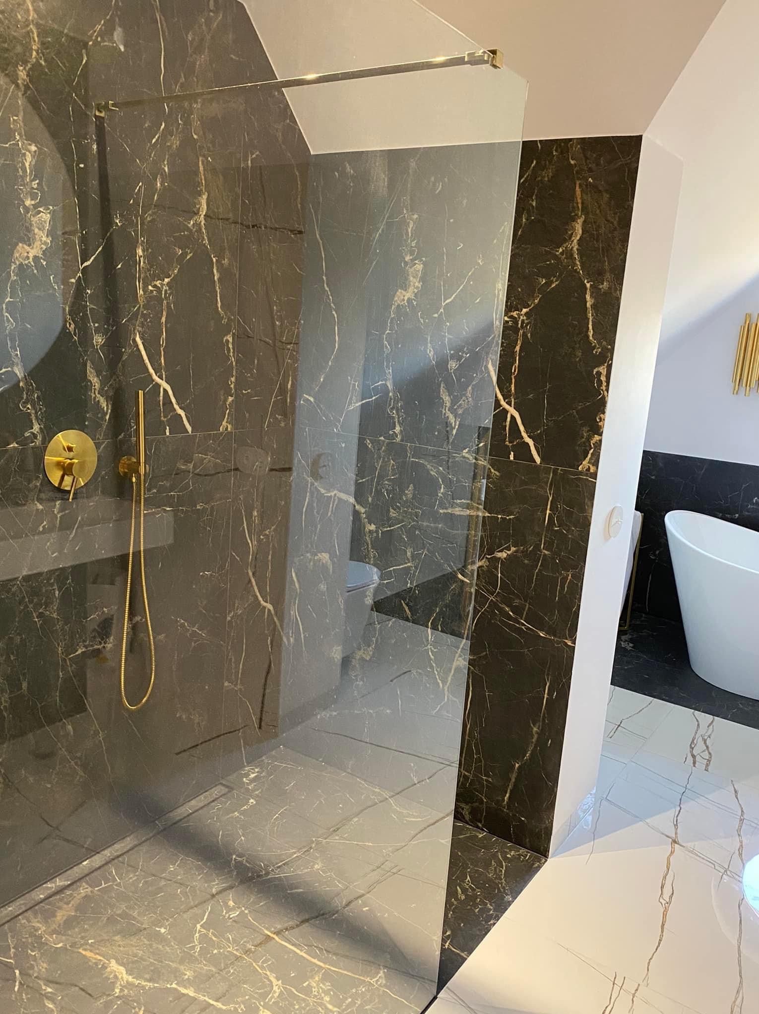 Nowoczesna łazienka z kabiną prysznicową typu walk-in, wykończona czarnym marmurem z złotymi żyłami, złotą armaturą i odpływem liniowym w podłodze, widoczna wanna wolnostojąca w tle.