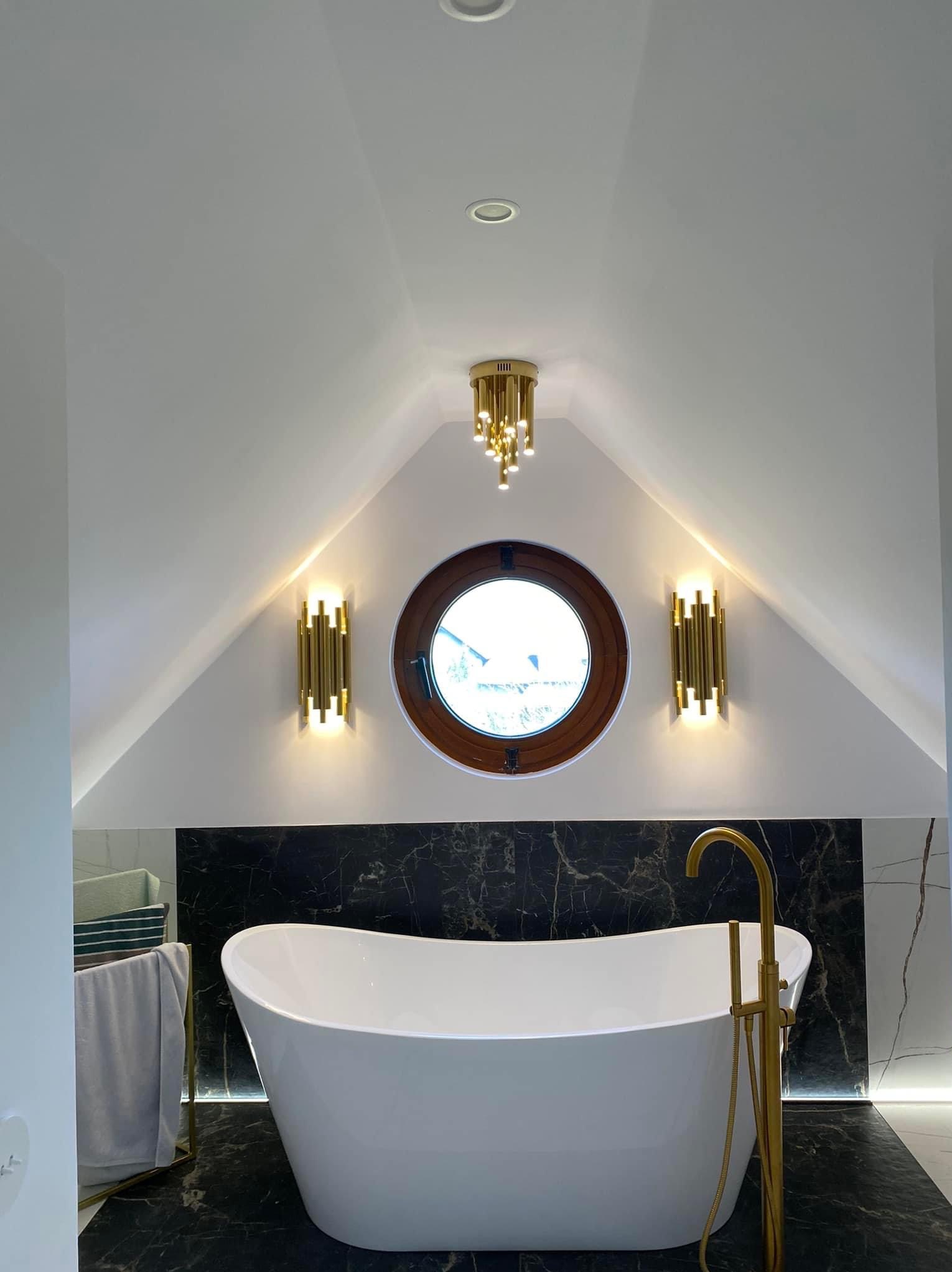 Nowoczesna łazienka na poddaszu z wolnostojącą białą wanną, złotą armaturą, okrągłym oknem i dekoracyjnym oświetleniem w postaci złotych kinkietów i żyrandola.
