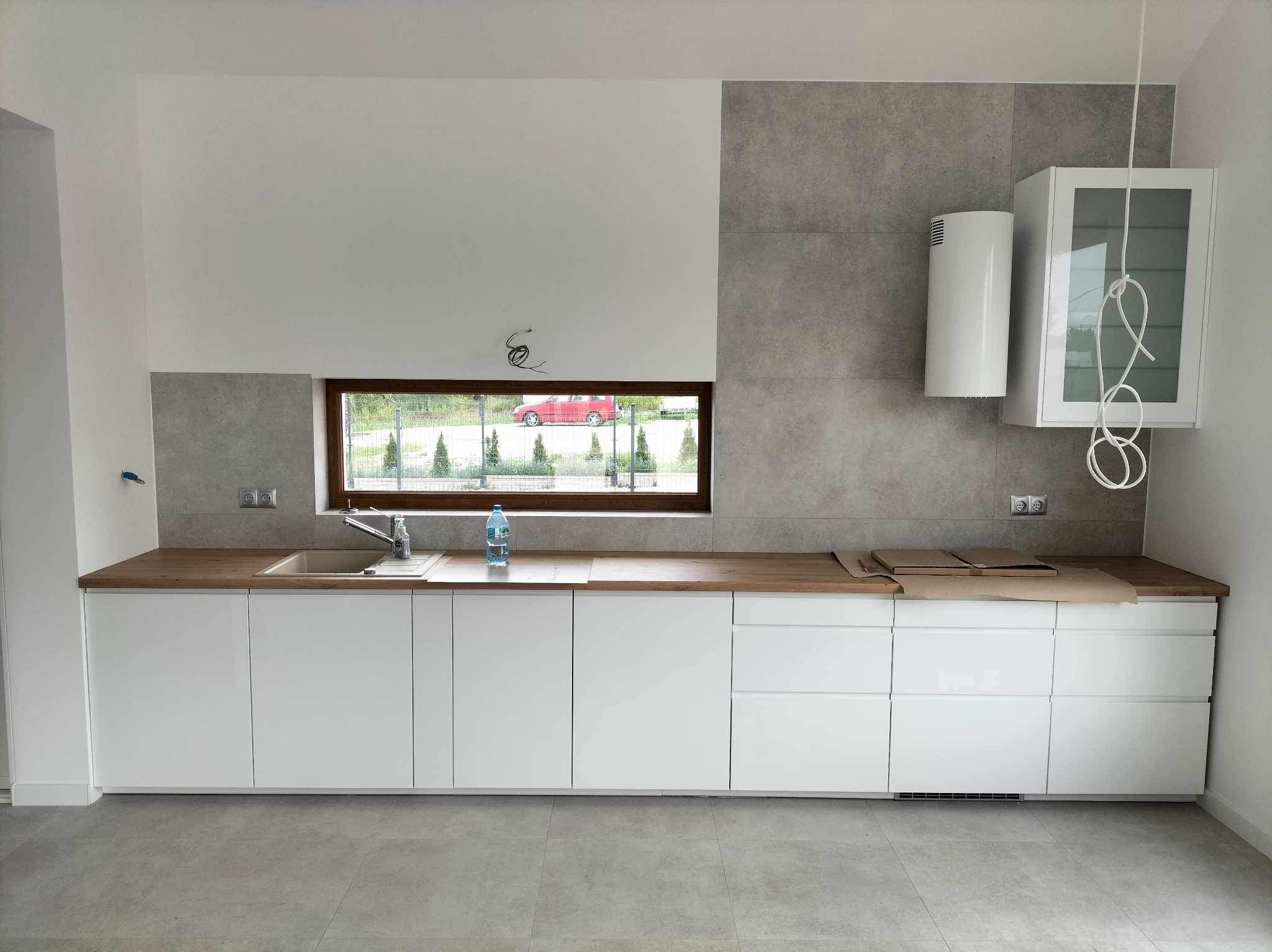 Nowoczesna kuchnia z białymi szafkami, drewnianym blatem, szarą płytką na ścianie, oknem z widokiem na ogród i wiszącym grzejnikiem.