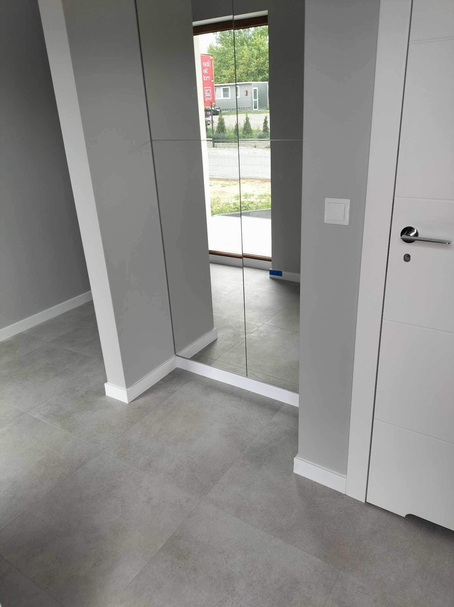 Nowoczesne wnętrze z szarą podłogą, lustrem w zabudowie i białymi drzwiami; widok na zewnątrz w odbiciu lustra.