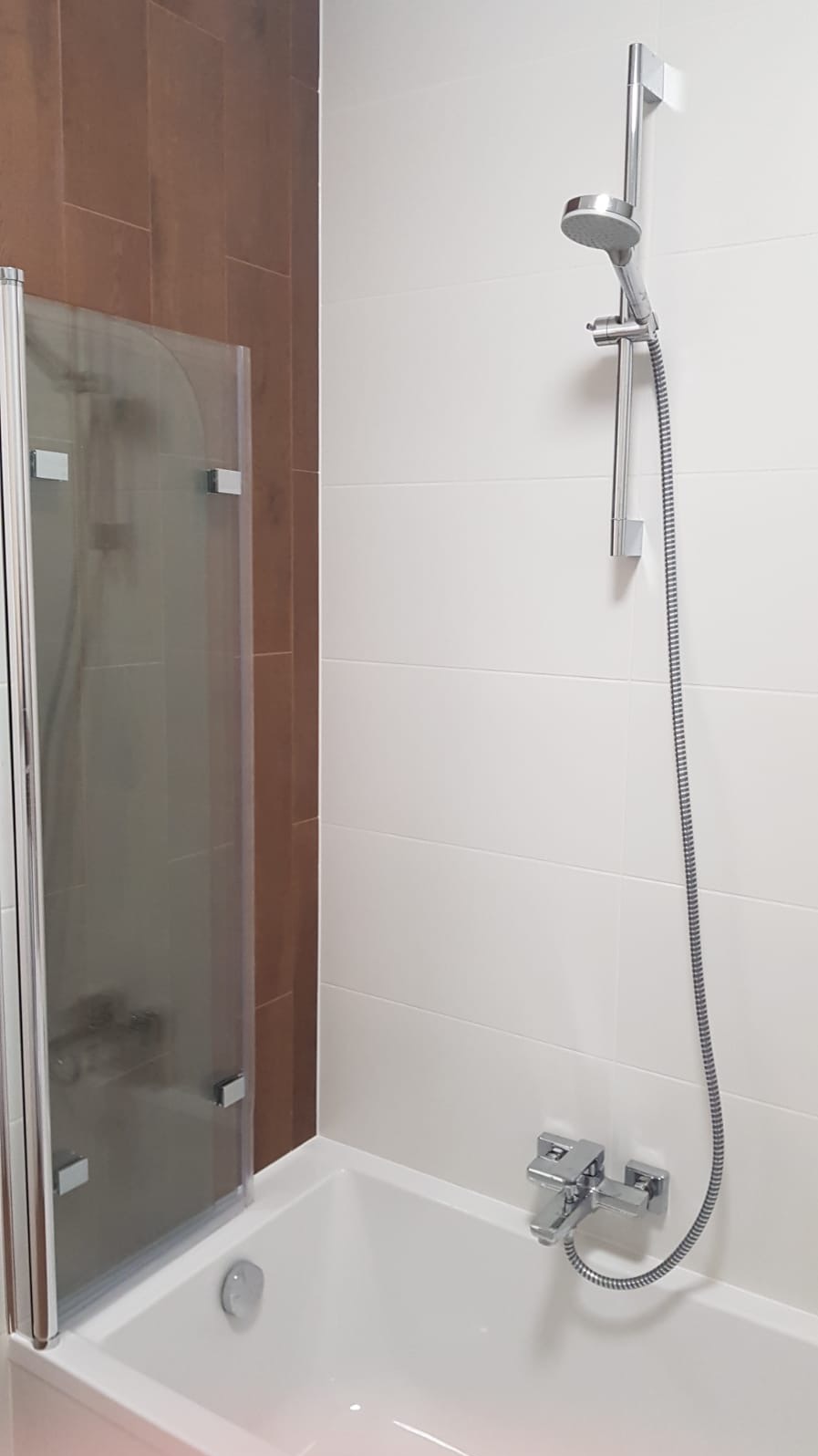 Nowoczesna łazienka z białą wanną, chromowaną armaturą i szklanym panelem prysznicowym na tle brązowych i białych płytek.