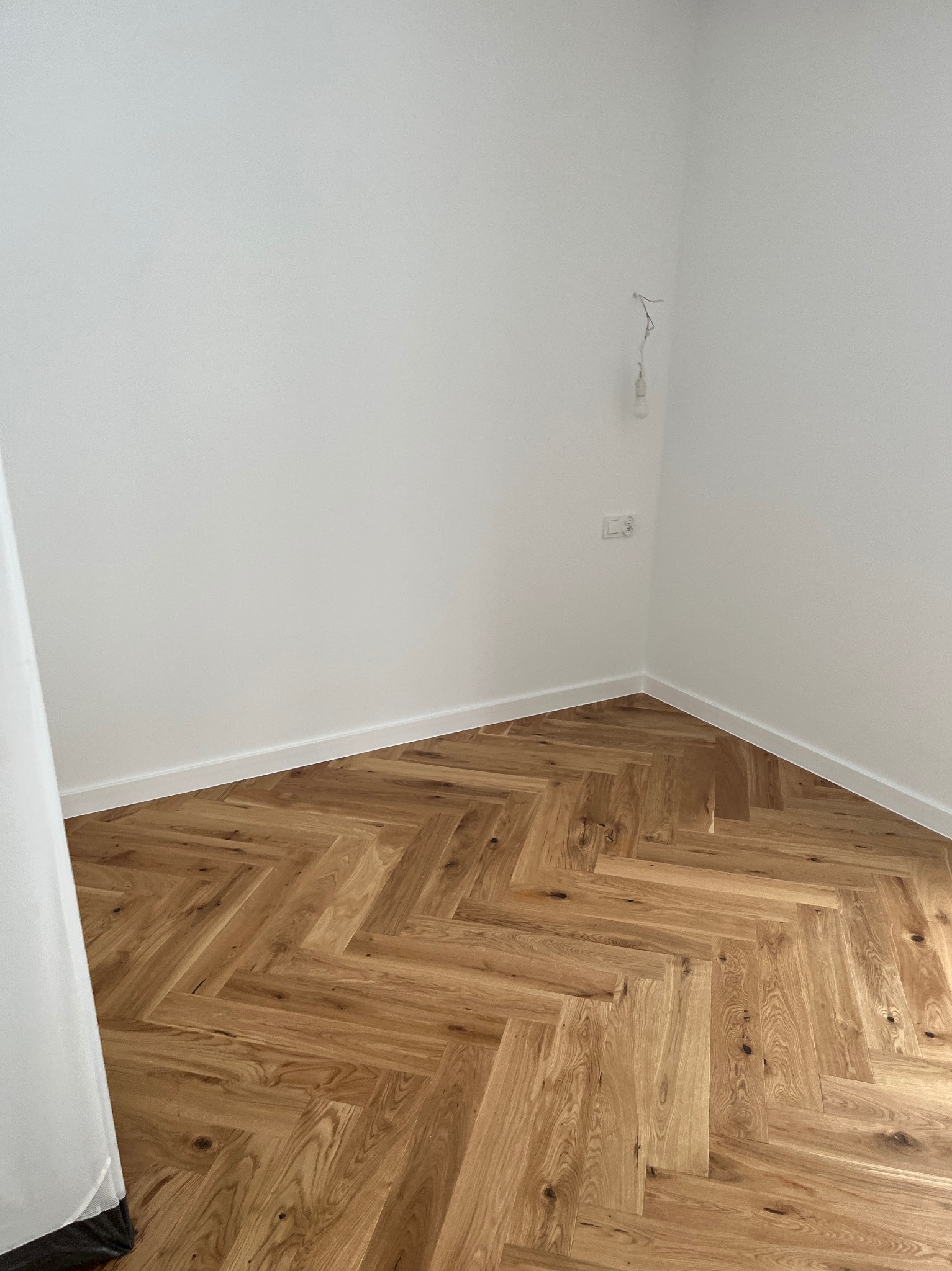 Ukończona podłoga z parkietu ułożonego w jodełkę w rogu pokoju z białymi ścianami i listwami przypodłogowymi. Widoczny fragment zasłony.