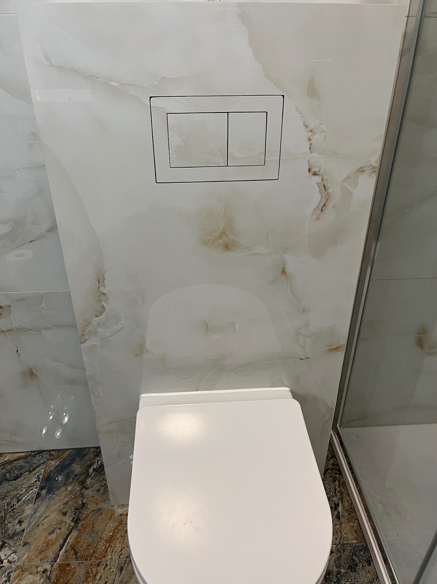 Wykończona łazienka: biała toaleta podwieszana z przyciskiem spłukującym na ścianie z płytek imitujących marmur, kabina prysznicowa z prawej strony.