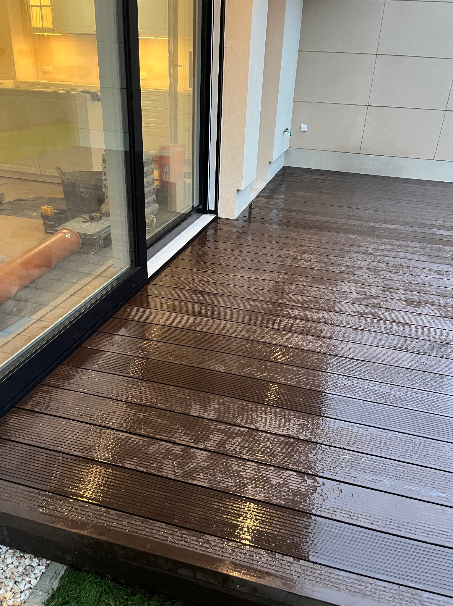 Nowoczesny taras z desek kompozytowych w odcieniach brązu, częściowo mokry, przylegający do budynku z dużym oknem. Widoczne narzędzia i materiały wewnątrz.