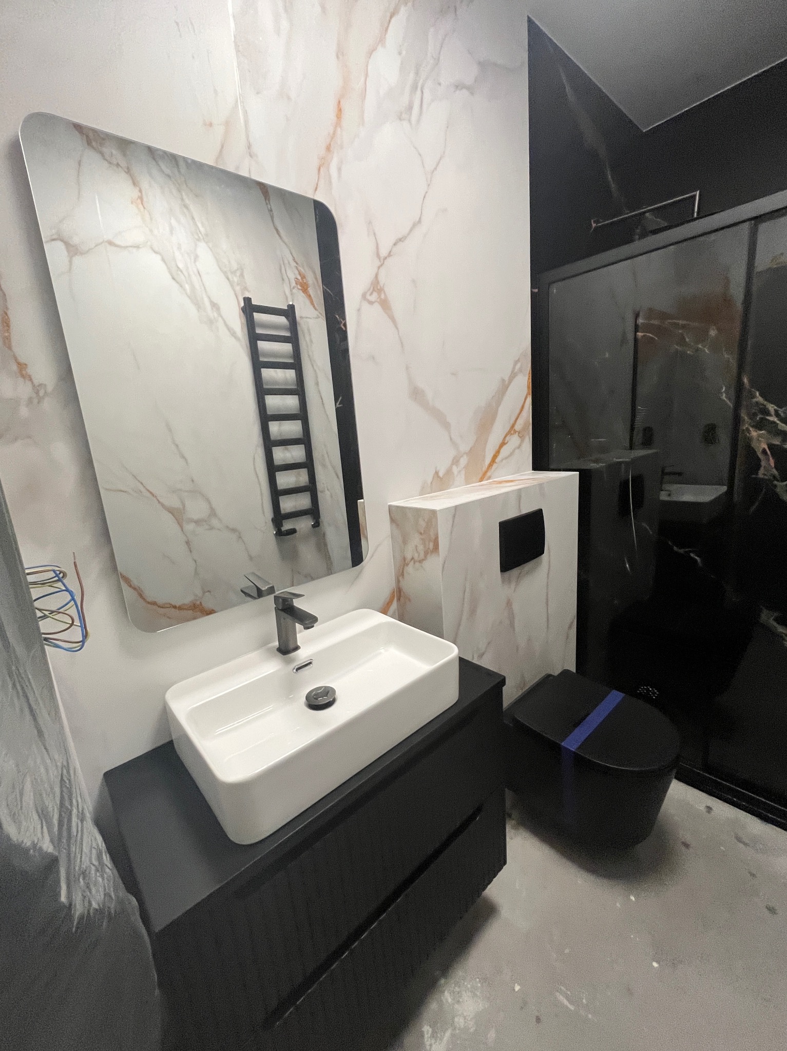 Nowoczesna łazienka z marmurowymi płytkami, czarną armaturą i podwieszaną toaletą z niebieskim akcentem. W lustrze odbija się grzejnik drabinkowy.