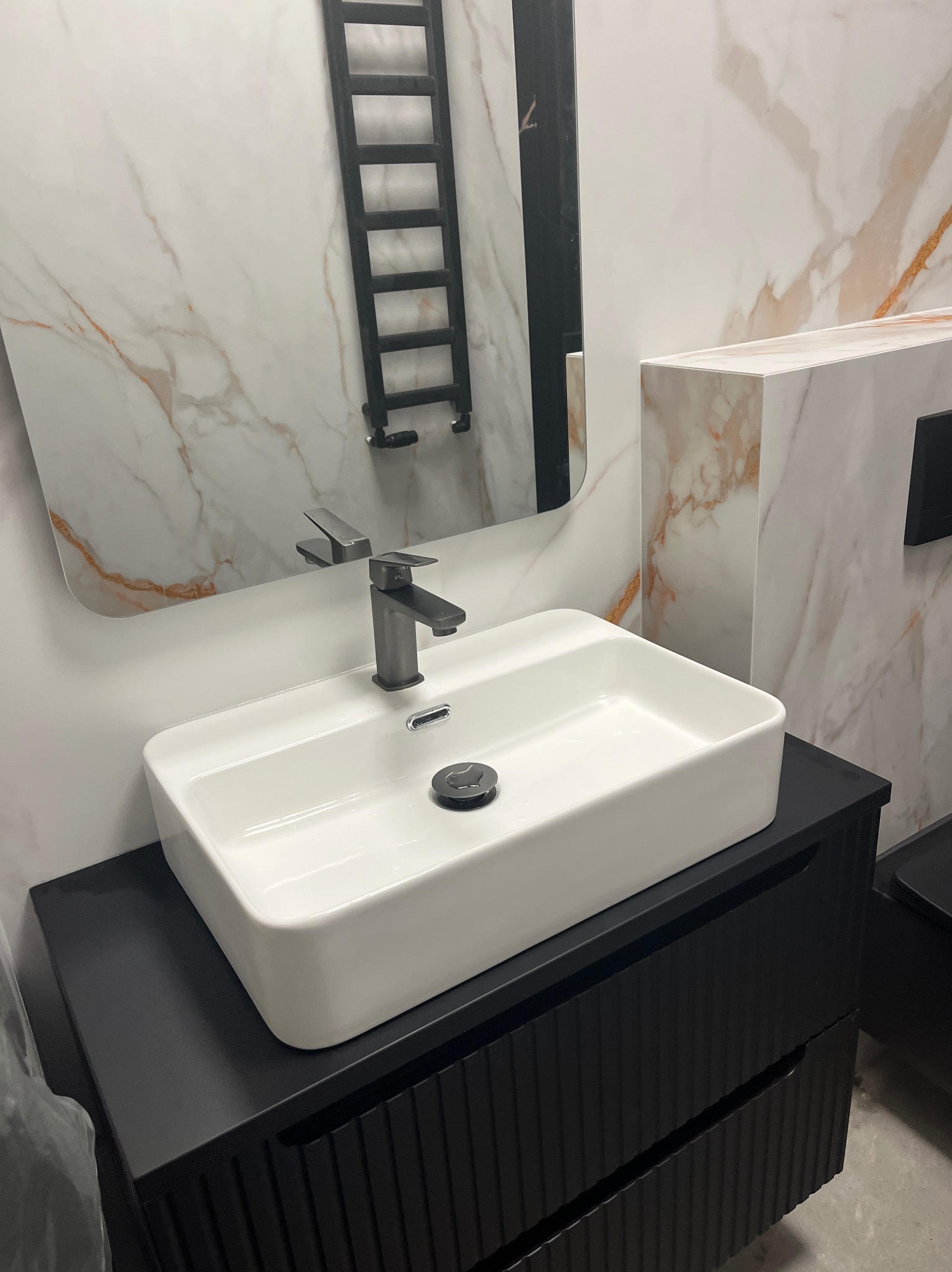 Nowoczesna łazienka: biała, prostokątna umywalka nablatowa z czarną szafką i baterią, marmurowe płytki na ścianie, grzejnik drabinkowy w lustrze.