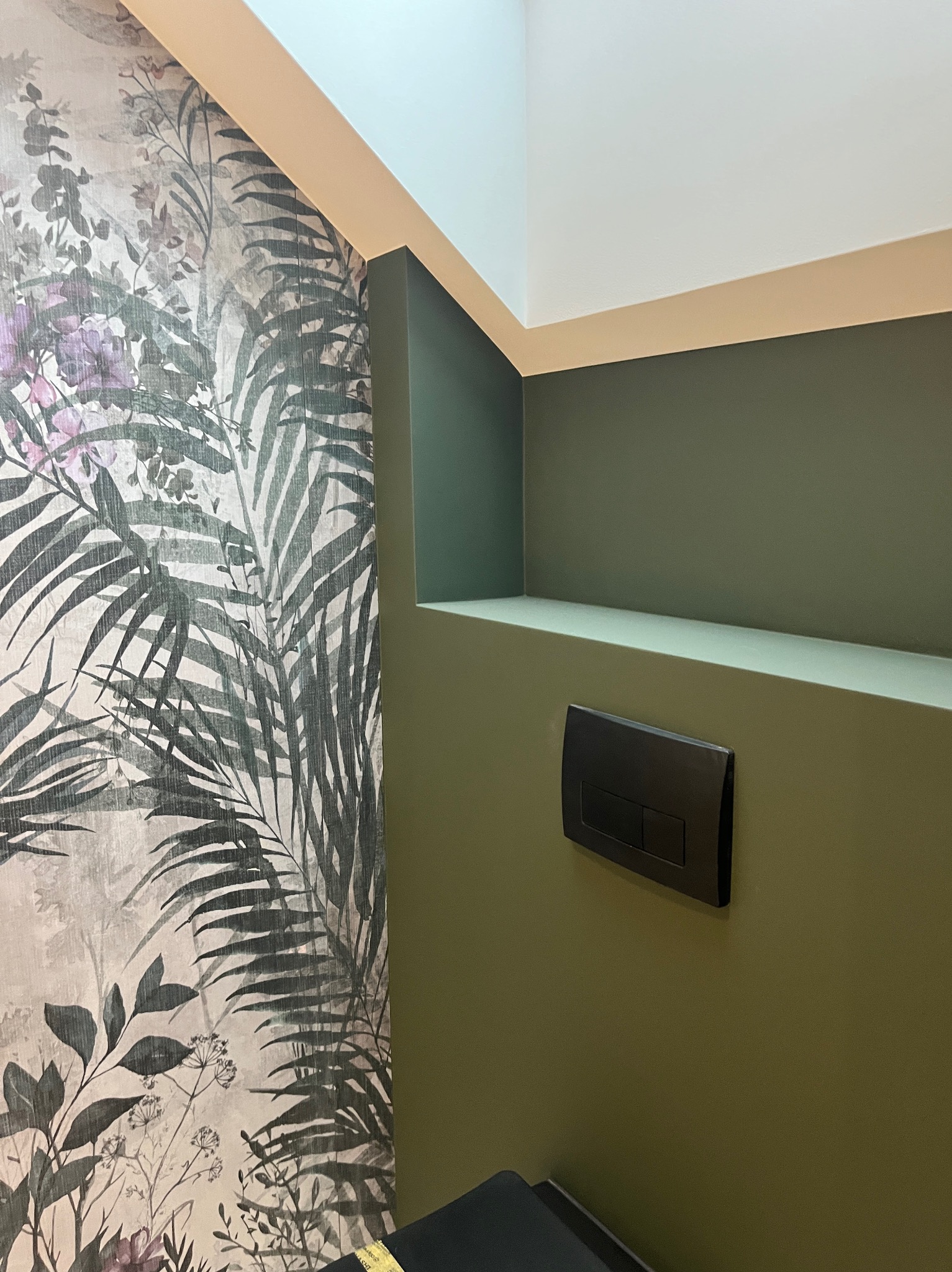 Wykończona łazienka: ściana z motywem roślinnym, zielona ściana z czarnym przyciskiem spłuczki, biały sufit. Nowoczesny design z akcentem natury.
