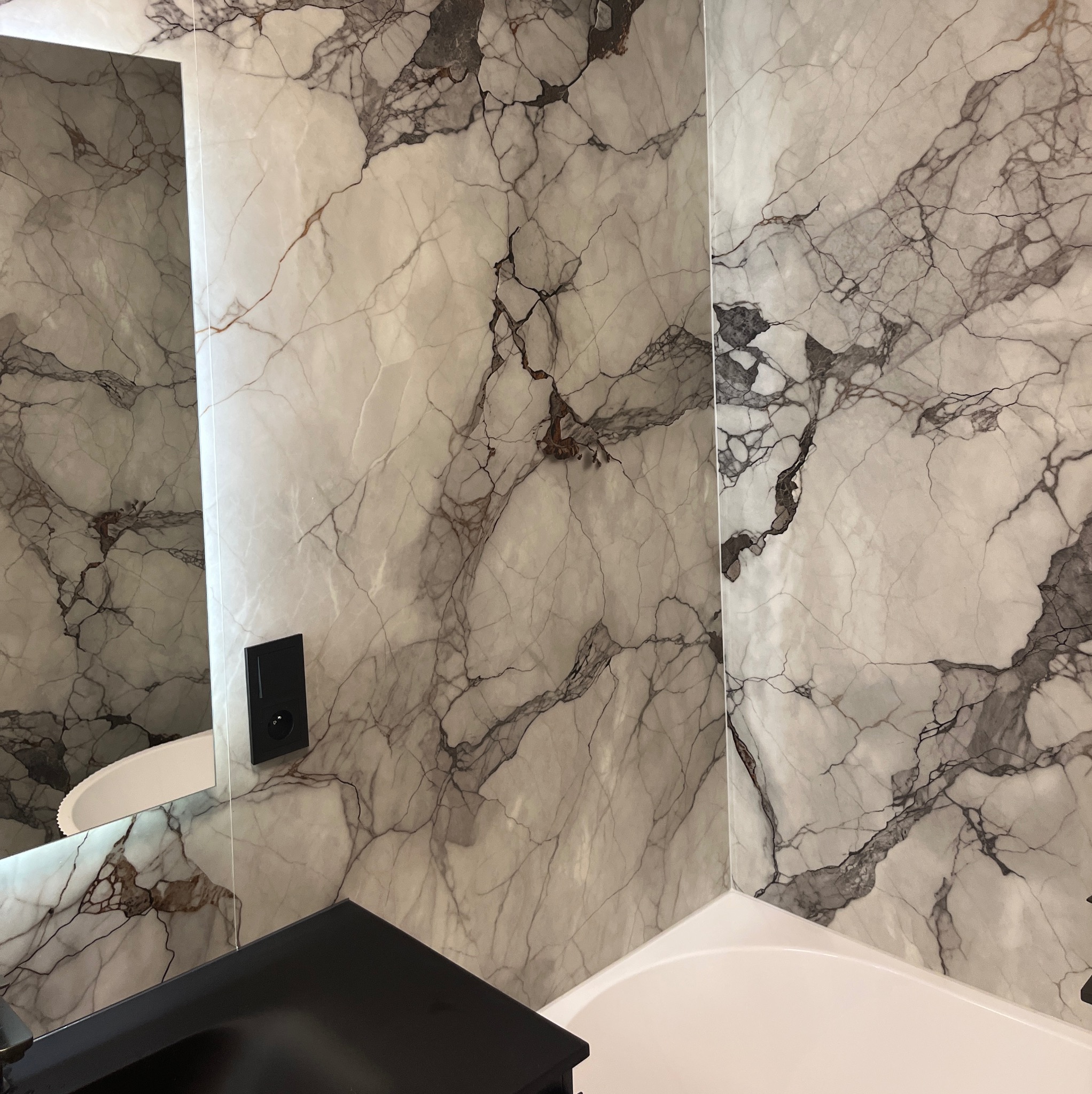 Wykończona łazienka z panelami ściennymi imitującymi marmur, czarna szafka pod umywalką i fragment wanny. Widoczne gniazdko elektryczne.