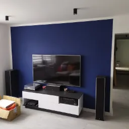 Pomieszczenie po malowaniu ściany na intensywny granat, z widocznym sprzętem audio-wideo i kartonem z akcesoriami, oświetlone punktowymi lampami sufitowymi.