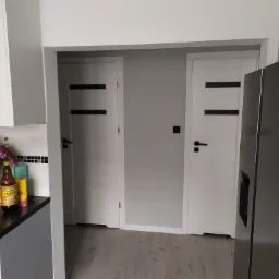 Nowoczesna kuchnia z dwiema białymi drzwiami z czarnymi paskami, szarą lodówką i blatem z kwiatami i butelkami, podłoga imitująca drewno.