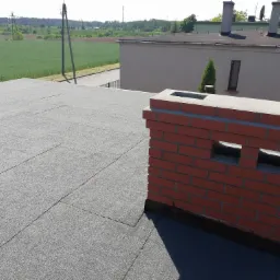 Szary dach pokryty papą z ceglanym kominem z otworami wentylacyjnymi, w tle budynek i zielone pole.