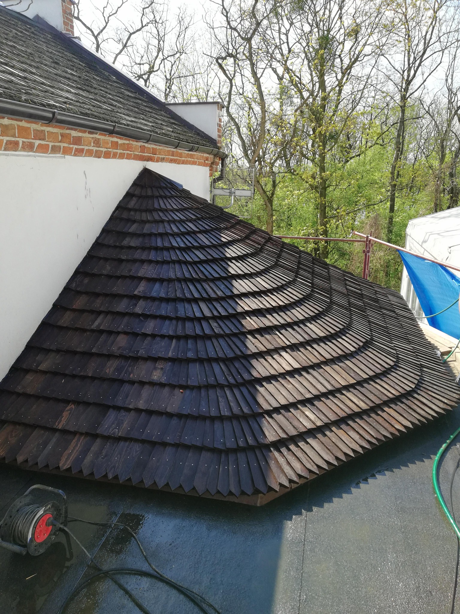 Stożkowy daszek pokryty ciemnym, drewnianym gontem, przylegający do białej ściany budynku z ceglanym wykończeniem, widok z góry, na dachu zwinięty przedłużacz i fragment węża ogrodowego.