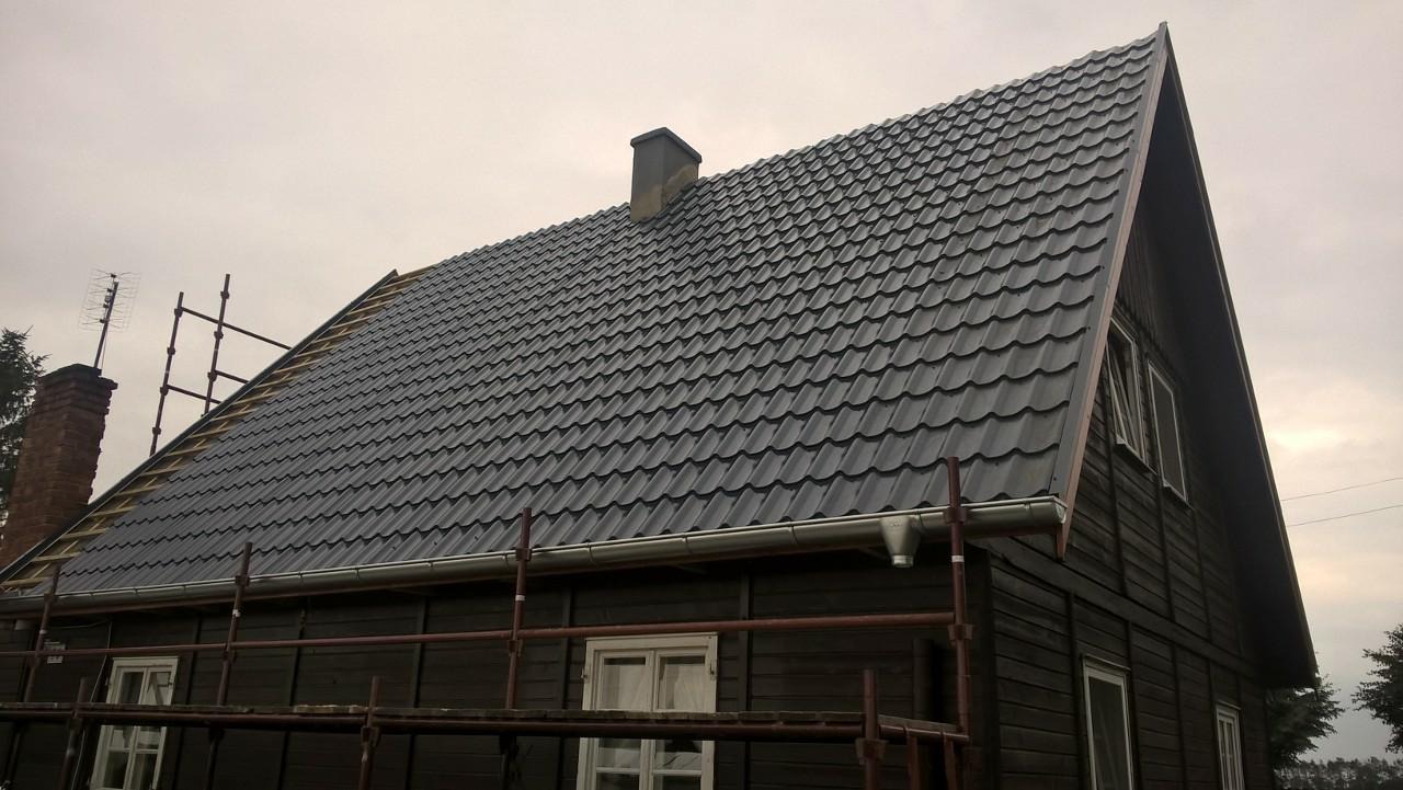 Ciemnoszary dach z blachodachówki na drewnianym domu, widoczne rusztowania i częściowo zamontowane rynny, komin z anteną.