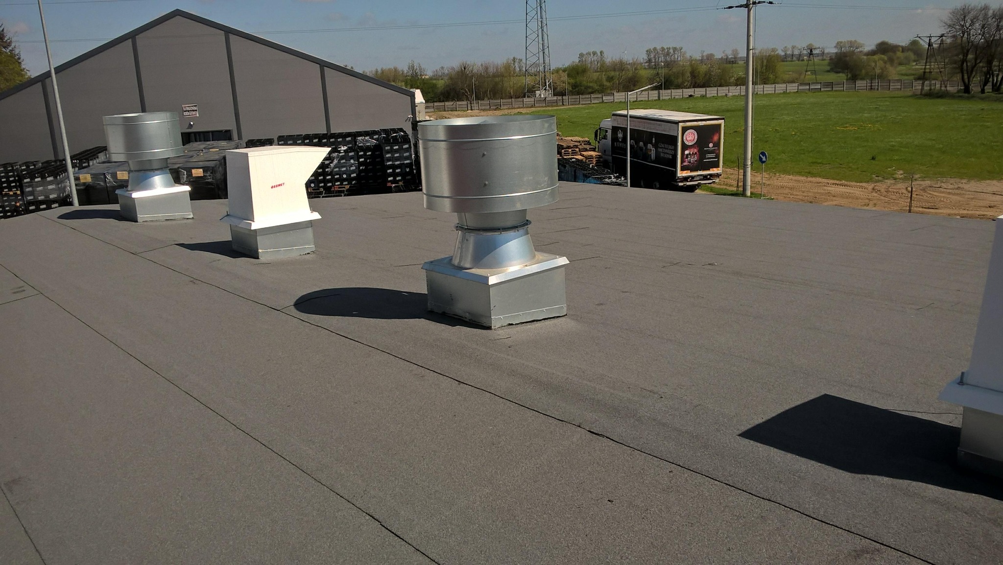 Szary, płaski dach przemysłowy z widocznymi metalowymi kominami wentylacyjnymi i elementami wentylacyjnymi, w tle budynek magazynowy, trawa i zaparkowana ciężarówka.