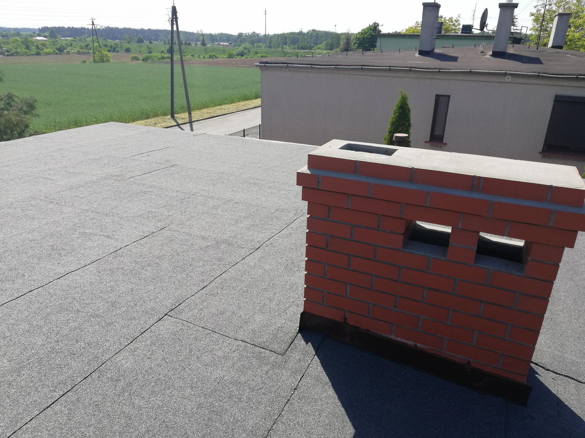 Szary dach pokryty papą z ceglanym kominem z otworami wentylacyjnymi, w tle budynek i zielone pole.