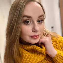 Portret młodej kobiety z delikatnym makijażem, błyszczącymi ustami i subtelnym cieniem do powiek, ubrana w żółty sweter z grubym splotem.