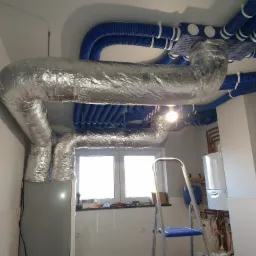 Srebrna, izolowana rura wentylacyjna w kształcie litery U, połączona z niebieskimi, karbowanymi przewodami wentylacyjnymi pod sufitem, widok na okno i drabinę.