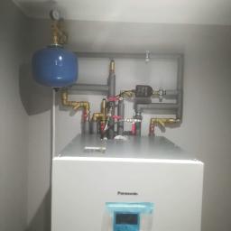 Usługi hydrauliczne - Biały piec Panasonic Aquarea z niebieskim zbiornikiem wyrównawczym i instalacją hydrauliczną z zaworami, rurami miedzianymi i szarymi rurami PCV.