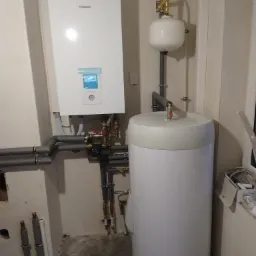 Biały piec dwufunkcyjny Panasonic Aquarea, zbiornik buforowy i rury grzewcze w narożniku pomieszczenia, widoczne elementy instalacji CO.