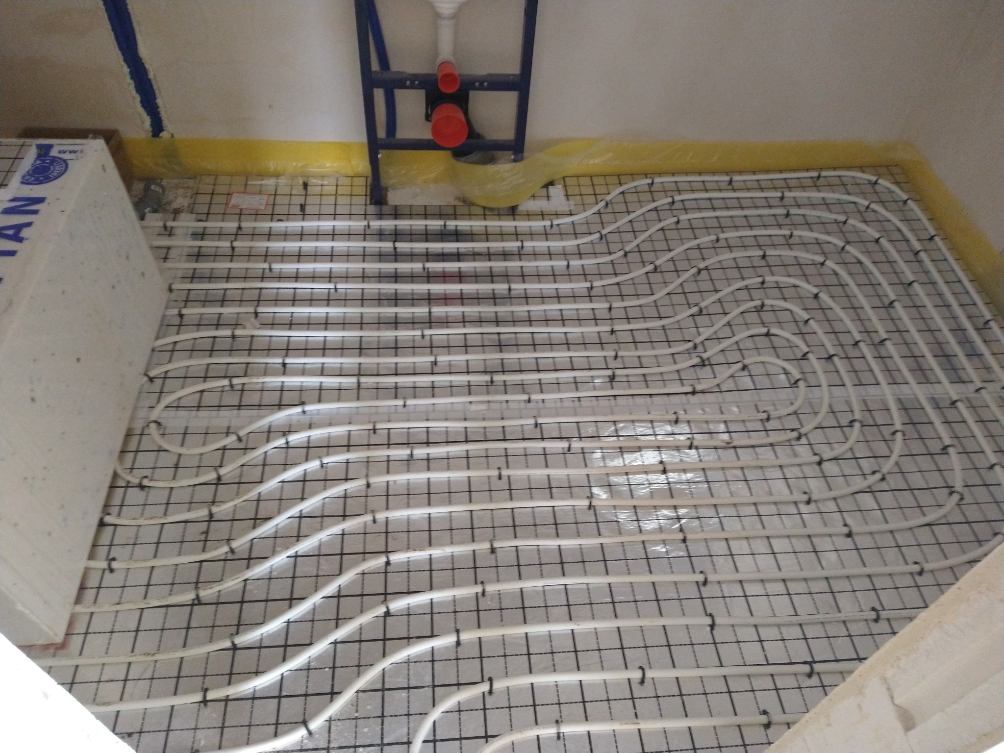 Białe rury ogrzewania podłogowego zamocowane do czarnej siatki na folii izolacyjnej, widok z góry, instalacja w trakcie montażu, element stelaża podtynkowego WC w tle.