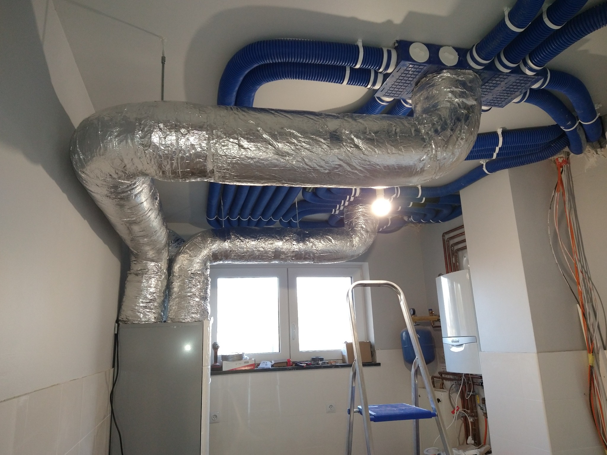 Srebrna, izolowana rura wentylacyjna w kształcie litery U, połączona z niebieskimi, karbowanymi przewodami wentylacyjnymi pod sufitem, widok na okno i drabinę.