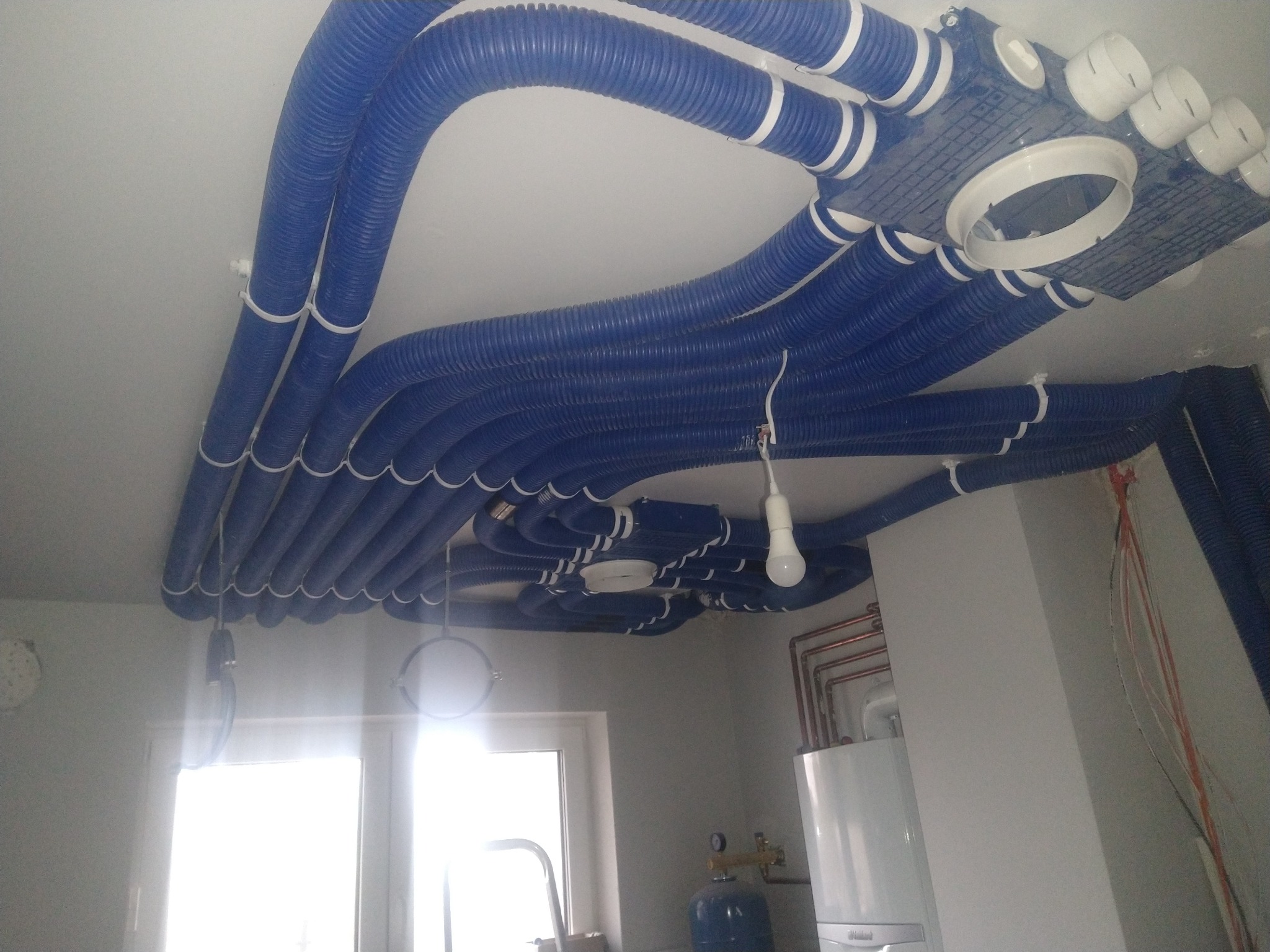 Niebieskie, karbowane rury wentylacyjne z białymi opaskami mocowane do sufitu, z widocznym rozdzielaczem i fragmentem instalacji grzewczej z piecem gazowym w tle.