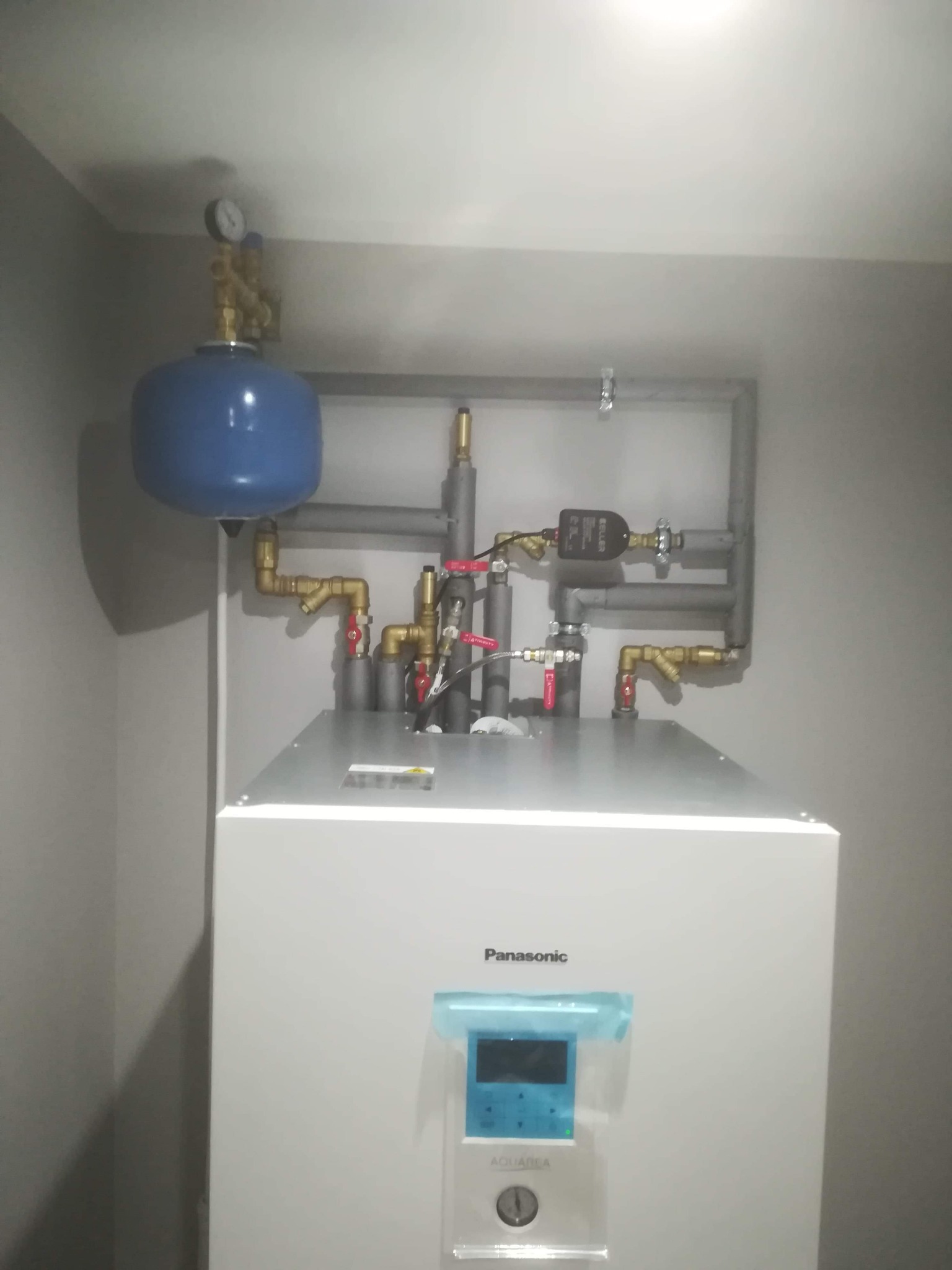Biały piec Panasonic Aquarea z niebieskim zbiornikiem wyrównawczym i instalacją hydrauliczną z zaworami, rurami miedzianymi i szarymi rurami PCV.