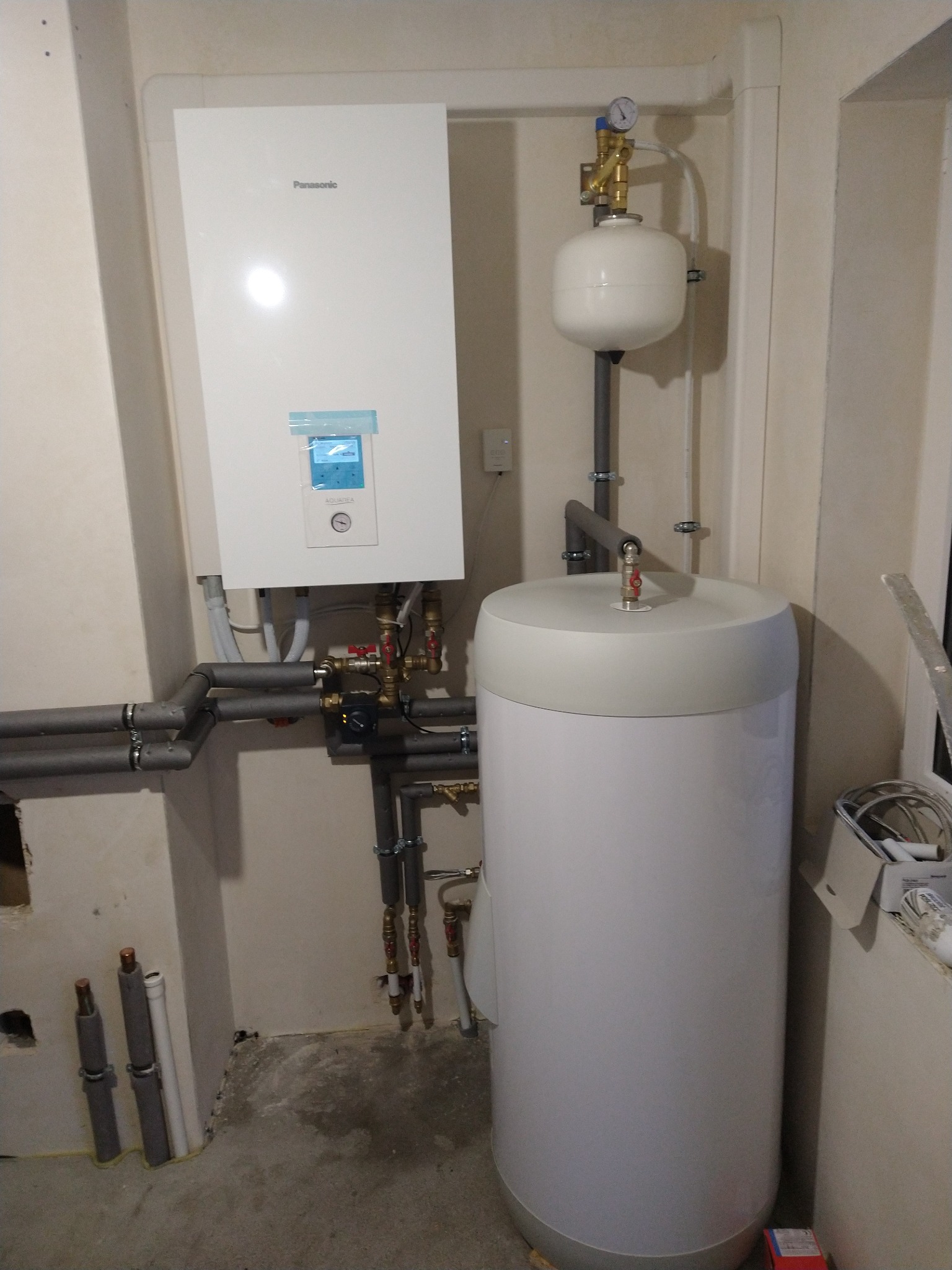 Biały piec dwufunkcyjny Panasonic Aquarea, zbiornik buforowy i rury grzewcze w narożniku pomieszczenia, widoczne elementy instalacji CO.