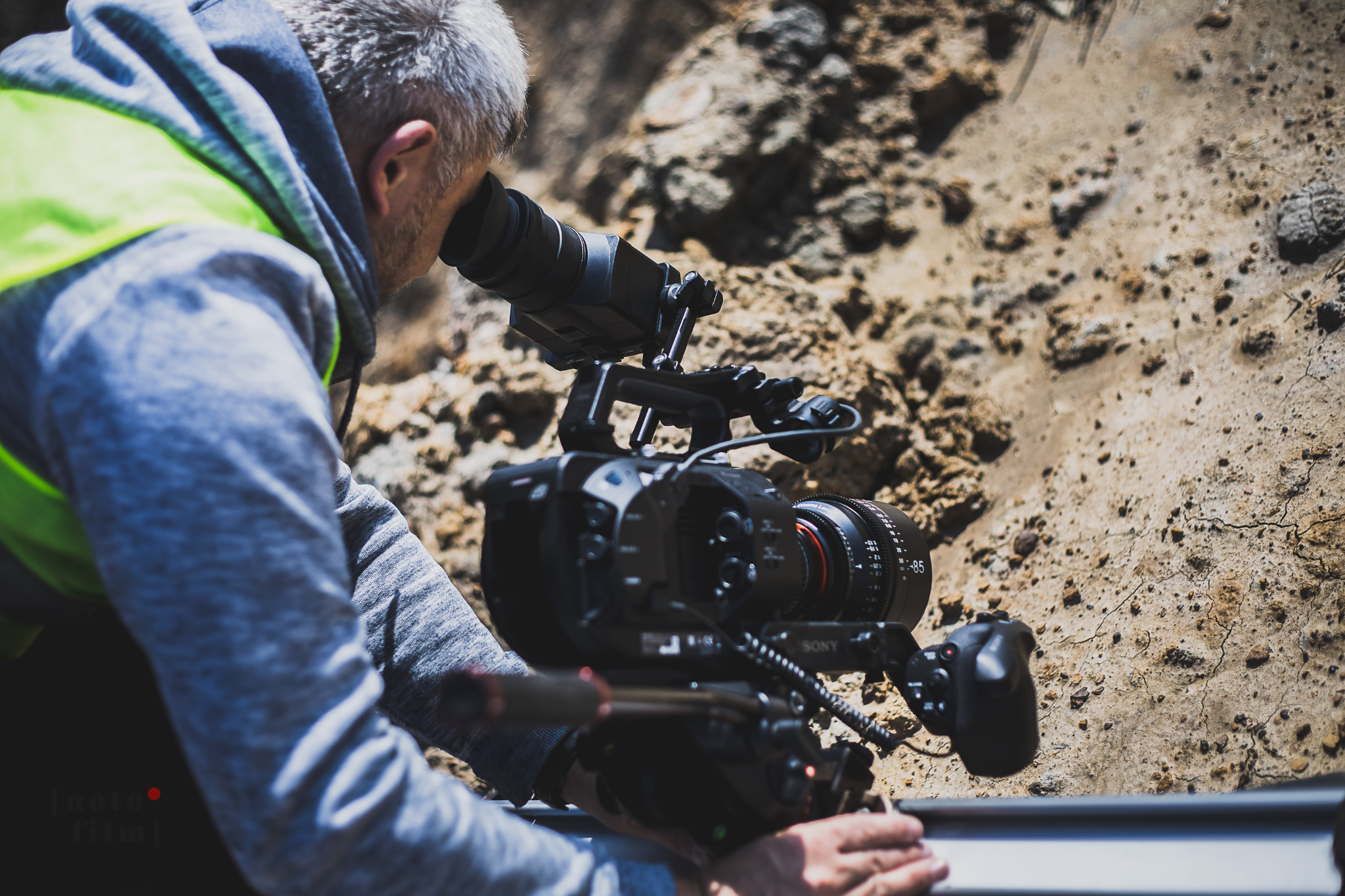Operator z siwymi włosami i w kamizelce odblaskowej obsługuje profesjonalną kamerę Sony na tle piaszczystego terenu.