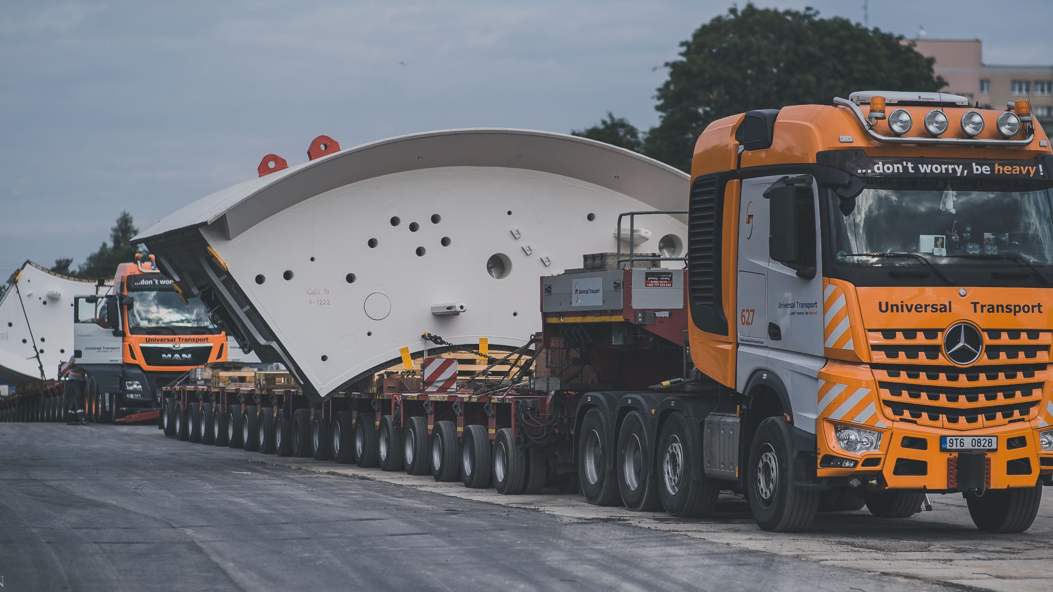 Dwa pomarańczowo-szare zestawy transportowe Universal Transport przewożą segmenty tunelu na wieloosiowych platformach; napis na ciężarówce: '...don't worry, be heavy!'.