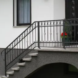 Czarne, metalowe balustrady schodowe z dekoracyjnymi elementami w kształcie skręconych prętów, prowadzące do wejścia budynku z donicą czerwonych kwiatów.