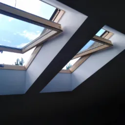 Dwa otwarte okna dachowe z drewnianymi ramami, wpuszczające światło dzienne i widok na błękitne niebo z pojedynczym drzewem, na tle białych ścian i ciemnego sufitu poddasza.