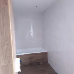 Wykończona łazienka z białymi płytkami ściennymi i panelami drewnopodobnymi na podłodze i obudowie wanny, widoczne otwory na oświetlenie w suficie.