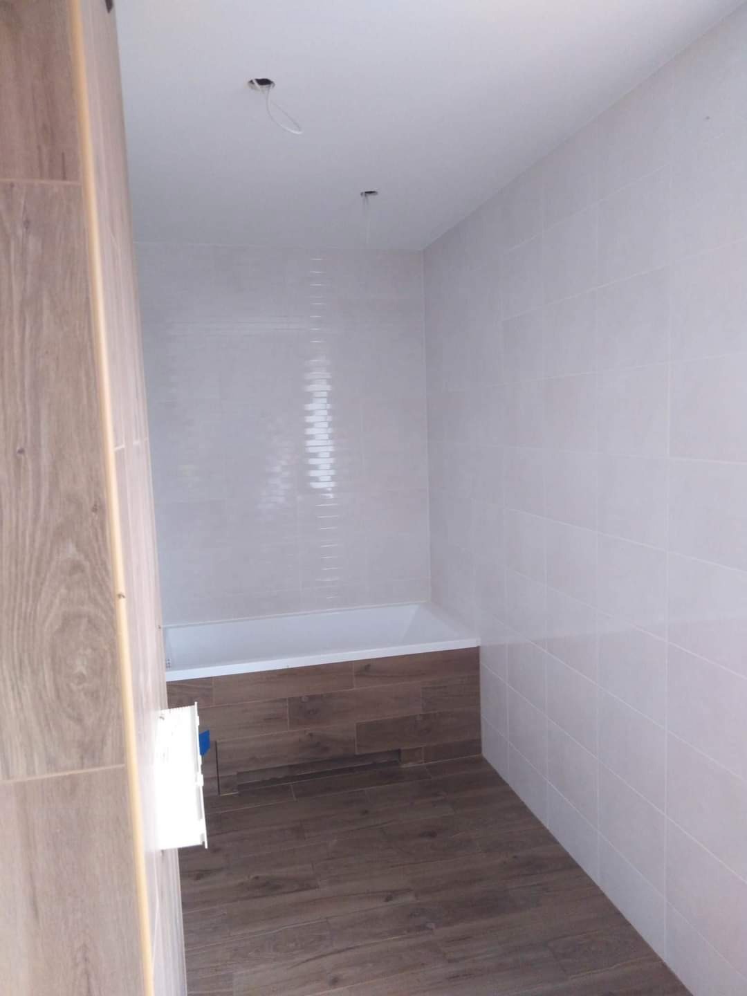 Wykończona łazienka z białymi płytkami ściennymi i panelami drewnopodobnymi na podłodze i obudowie wanny, widoczne otwory na oświetlenie w suficie.