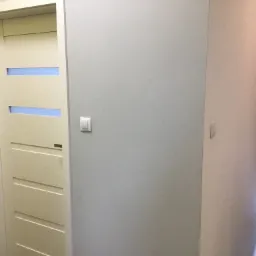 Białe drzwi wewnętrzne z poziomymi przeszkleniami w kolorze niebieskim, widoczne na tle jasnoszarej ściany z włącznikiem.