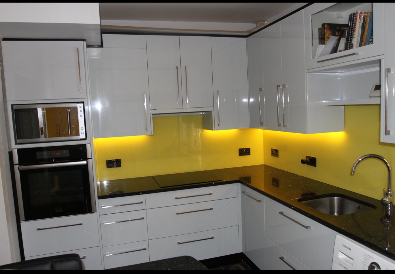 Nowoczesna kuchnia z białymi, lakierowanymi frontami, czarnym blatem i żółtym panelem szklanym podświetlonym LED, z widocznym zlewem i sprzętem AGD.