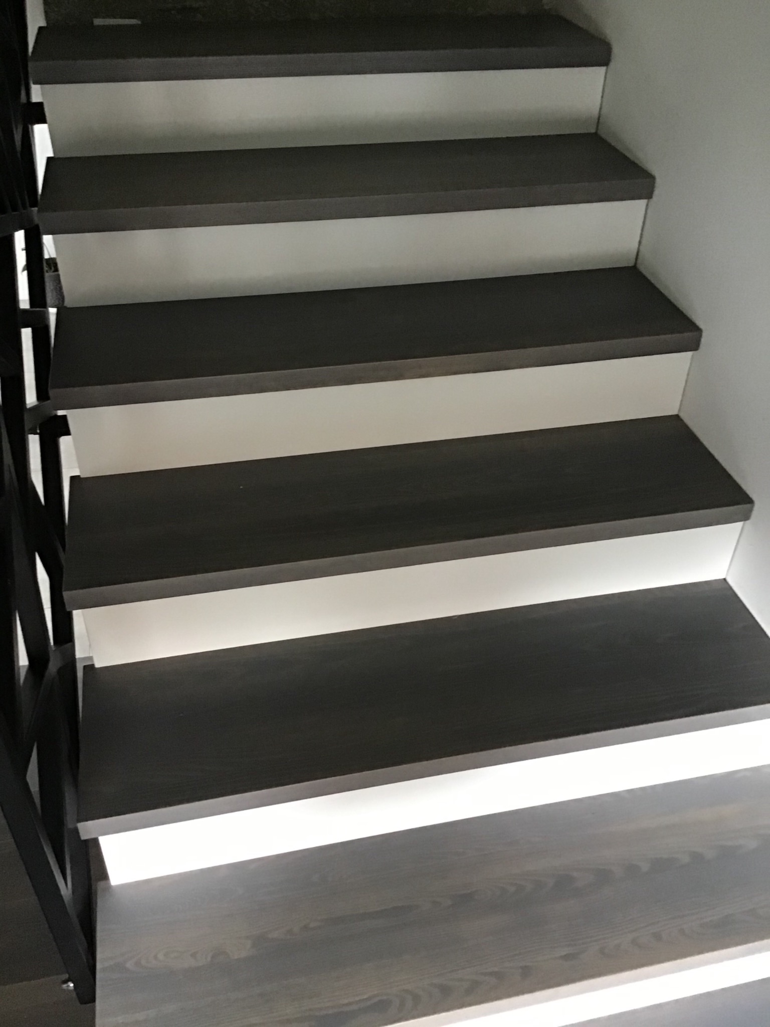 Nowoczesne schody z ciemnego drewna z podświetleniem LED pod każdym stopniem, widoczne metalowe elementy balustrady, minimalistyczny design.
