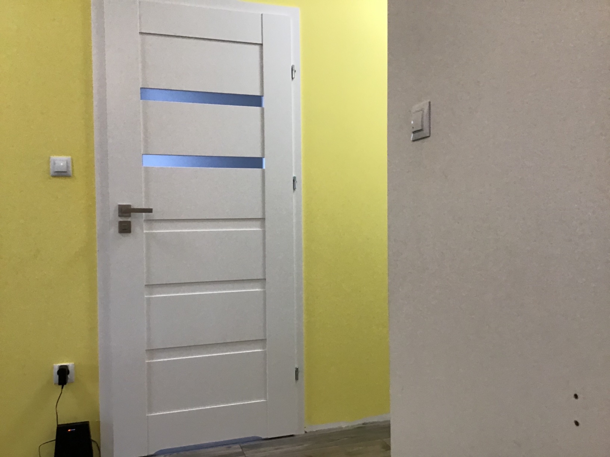 Białe drzwi wewnętrzne z poziomymi przeszkleniami na tle żółtej ściany, widoczny włącznik światła i gniazdko elektryczne.