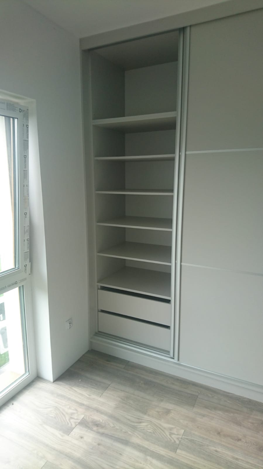 Biała szafa wnękowa z przesuwnymi drzwiami, widoczne półki i dwie szuflady, jasne panele podłogowe, ujęcie przy oknie.