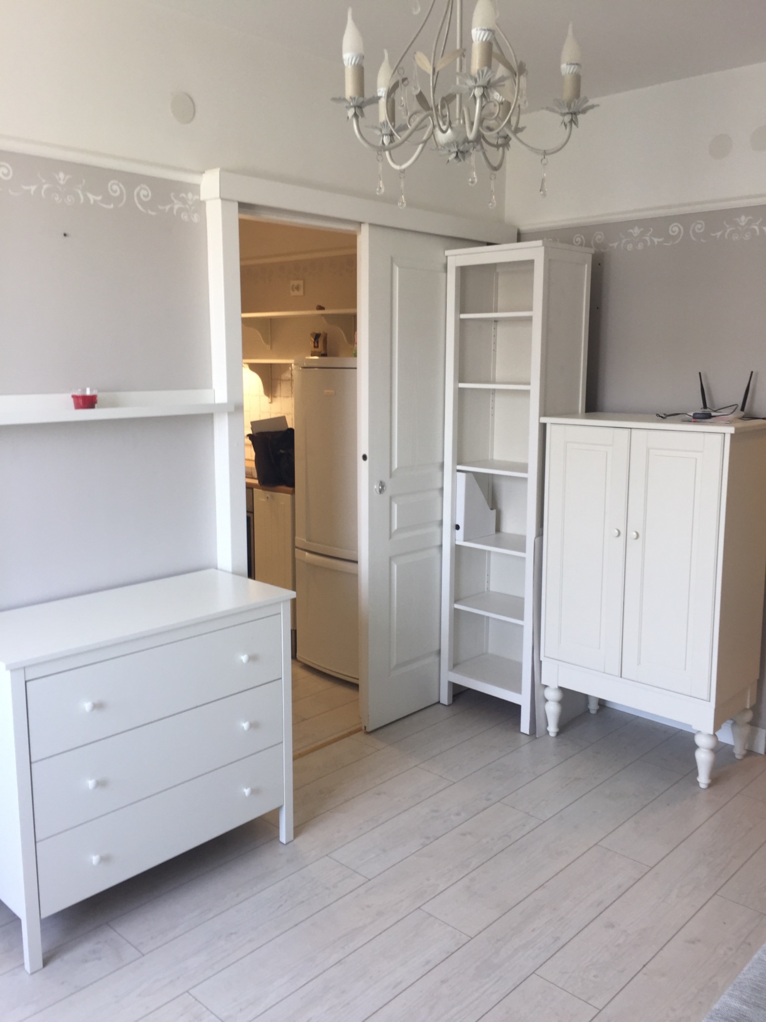 Jasne wnętrze z białymi meblami: komoda z trzema szufladami, regał, szafka z dwojgiem drzwi, widok na kuchnię przez otwarte drzwi przesuwne, dekoracyjny wzór na ścianie.