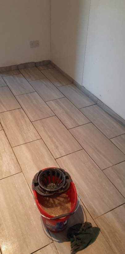 Ułożone płytki podłogowe imitujące drewno w rogu pomieszczenia, widoczne gniazdko elektryczne na ścianie, wiadro z mopem i ścierka na podłodze.