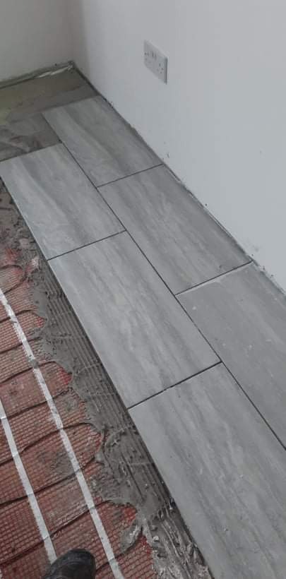 Układanie szarych, podłużnych płytek na podłodze z ogrzewaniem podłogowym, widoczny klej i fragment buta roboczego w rogu.