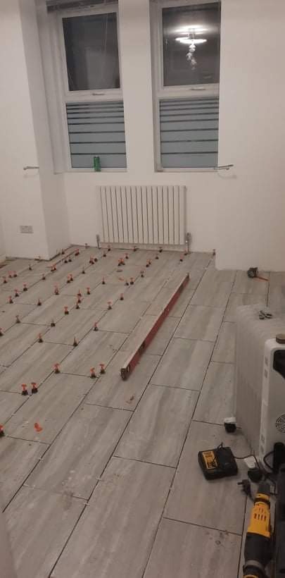 Układanie szarych płytek podłogowych z użyciem systemu poziomowania płytek w remontowanym pomieszczeniu. Widoczny grzejnik, okna z żaluzjami i narzędzia pracy.