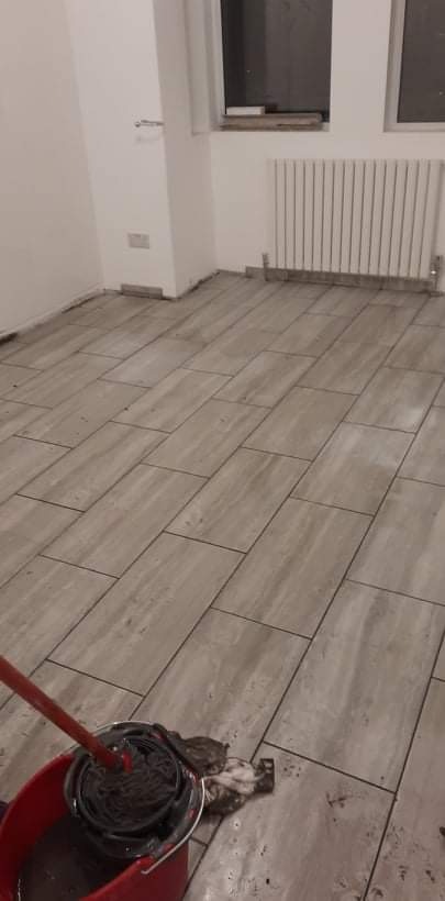 Nowo położone, szare płytki podłogowe w pokoju, czyszczone mopem zanurzonym w czerwonym wiadrze z brudną wodą; widoczny fragment ściany z grzejnikiem i oknem.